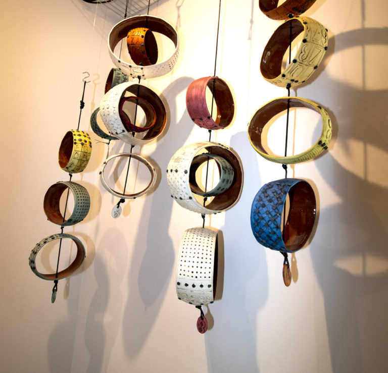 A series of pottery wall hanging art by Ann Ohotto Thompson. They are comprised of a series of hanging shapes, mostly round like bowls without bottoms. Thick black cord is tied between them, so a set of 5 or 6 hang down in a row. They are white, with an accent of orange and pink on the inner bowl shaped pieces. This pottery is by Ann Ohotto Thompson for her pottery shop, County 8 Pottery.