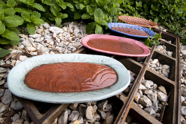 A line of four handmade serving trays sit on a metal grate outside by some green mint plants. They are all oval shaped ,and each has different textures and patterns carved into the top. They are all brown in the center, with a different color rim on each one, one is light blue, pink, dark blue, and orange. They are all handmade pottery by Ann Ohotto Thompson for her pottery shop, County 8 Pottery.