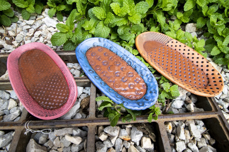 Three handmade serving trays sit outside on a metal grate by some green plants. They are all oval shaped, with different patterns on each one. The tray on the left is pink and brown, in the middle is blue and brown, and the right is orange and brown. All three are made by Ann Ohotto Thompson for her pottery shop, County 8 Pottery.