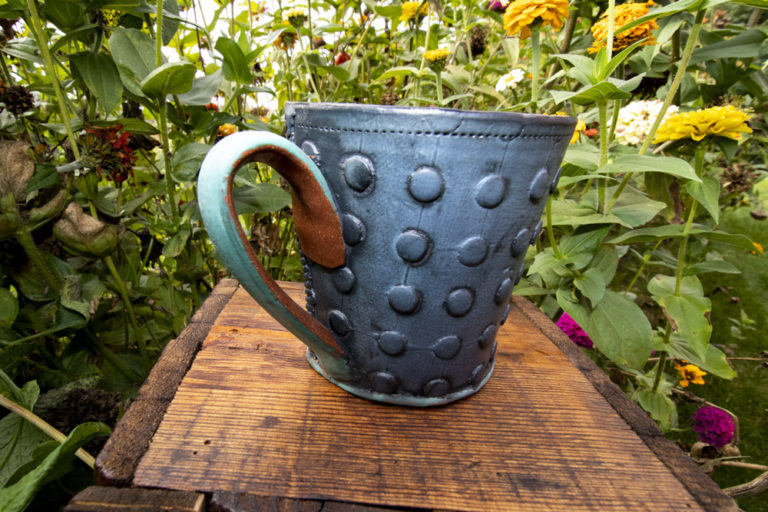 A dark teal, light blue, and brown mug sits on a dark wooden stand outside in front of green leaves and yellow flowers. The mug has artistic textures both inside and outside of the cup. This coffee mug is by Ann Ohotto Thompson for her pottery shop, County 8 Pottery.