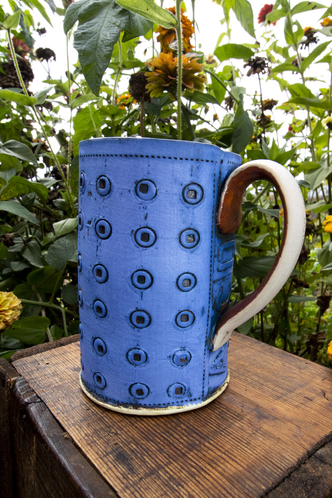 A bright blue and white mug sits on a dark wooden table in front of green leaves and flowers. The mug has artistic textures and patterns both inside and outside the cup. This piece is by Ann Ohotto Thompson for her pottery shop, County 8 Pottery.