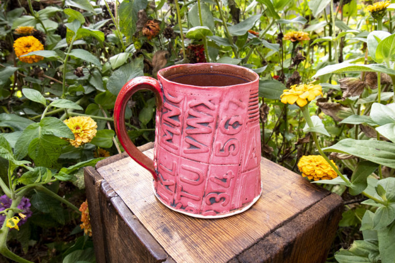 A red coffee mug sits on a little dark brown wooden stand, surrounded by green leaves and yellow flowers. The cup has texture and patterns on the cup, handle, and inside of the mug. The mug was handmade by Ann Ohotto Thompson for County 8 Pottery.