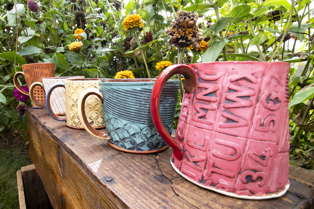 A group of five handmade coffee mugs sit displayed on a dark brown bench outside, in front of green leaves and yellow flowers. The cups are all different colors, blue, red, orange, and teal, and each has different interesting textures and patterns on them. They are handmade by Ann Ohotto Thompson for County 8 Pottery.