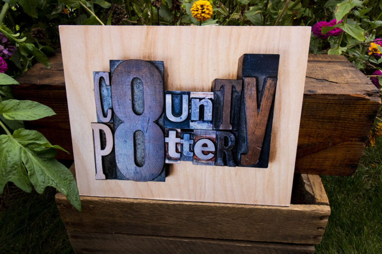 A wooden sign in front of a dark brown wooden bench, among green leaves and wildflowers. The sign reads, "County 8 Pottery". Each letter is a different color and size.