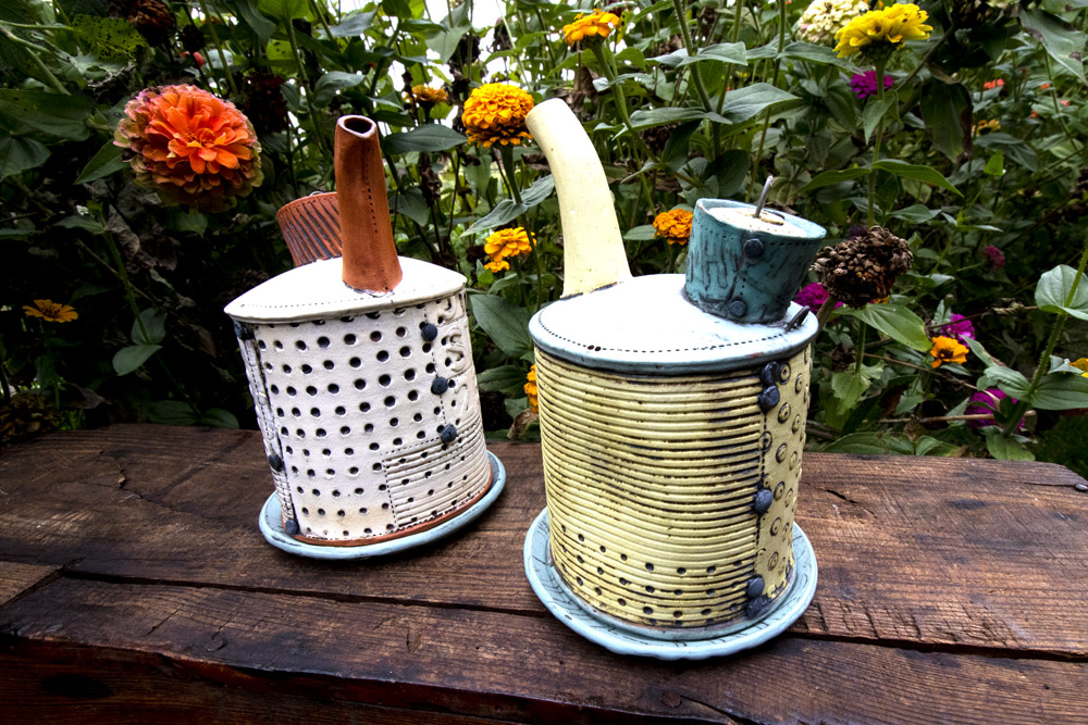 Two handmade pottery teapots sit on a dark brown wooden bench in front of a clump of yellow wildflowers. One teapot is white and orange, and the other is yellow and light blue. Both are made by Ann Ohotto Thompson for County 8 Pottery.