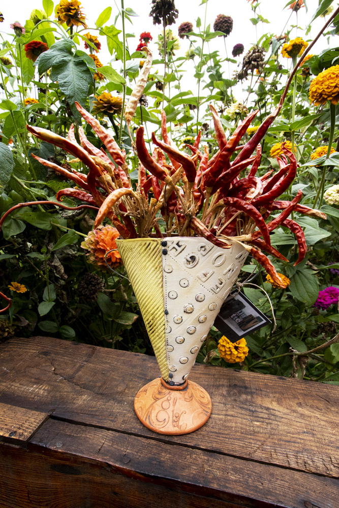 A yellow and white textured handmade pottery vase sits on a dark brown wooden bench. There are a bunch of decorative dried chilis in the vase. Behind the vase is a background of green leaves and yellow and orange wildflowers. The vase is by Ann Ohotto Thompson for County 8 Pottery.