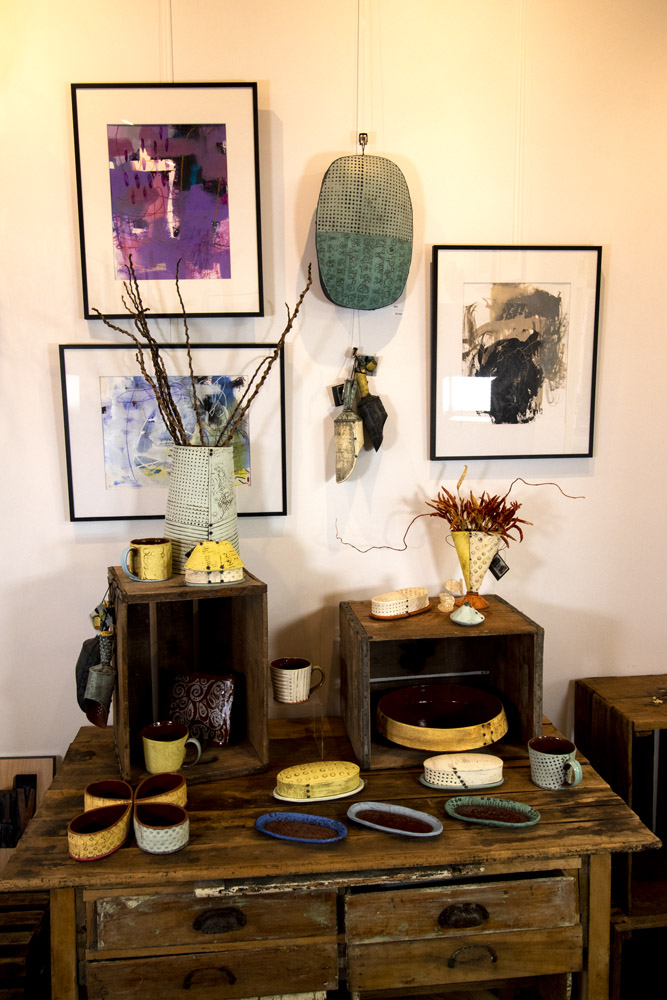 A display of Ann Ohotto Thompson's handmade pottery at County 8 Pottery. There is a dark wooden table with lots of decorative bowls and serving platters displayed on top, and covered butter dishes and vases with dried flowers are sitting on display pedestals on the table. On the wall behind the table are three framed abstract art prints, and a pottery art wall hanging by Ann Ohotto Thompson.