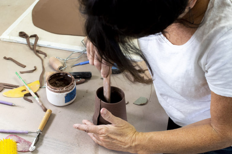 Ann Ohotto Thompson is shown from slightly above, so we can see what she is working on. She is forming a red clay coffee mug with her hands and a long pottery forming tool. Around her on the table are other pottery sculpting tools, paintbrushes, cups, and long scraps of clay.