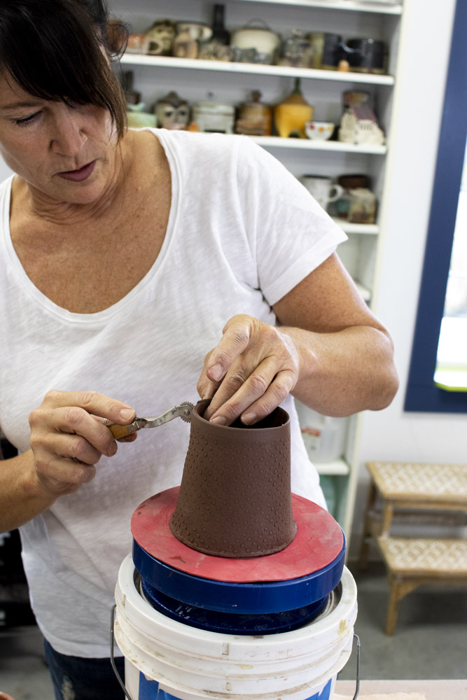 Ann Ohotto Thompson is shown working a red clay coffee mug with a pottery texturizing tool. She is wearing a white t-shirt and looking intently at her work. Behind her are white shelves filled with colorful pottery creations, mugs, and sculptures. She is inside her pottery studio at County 8 pottery.