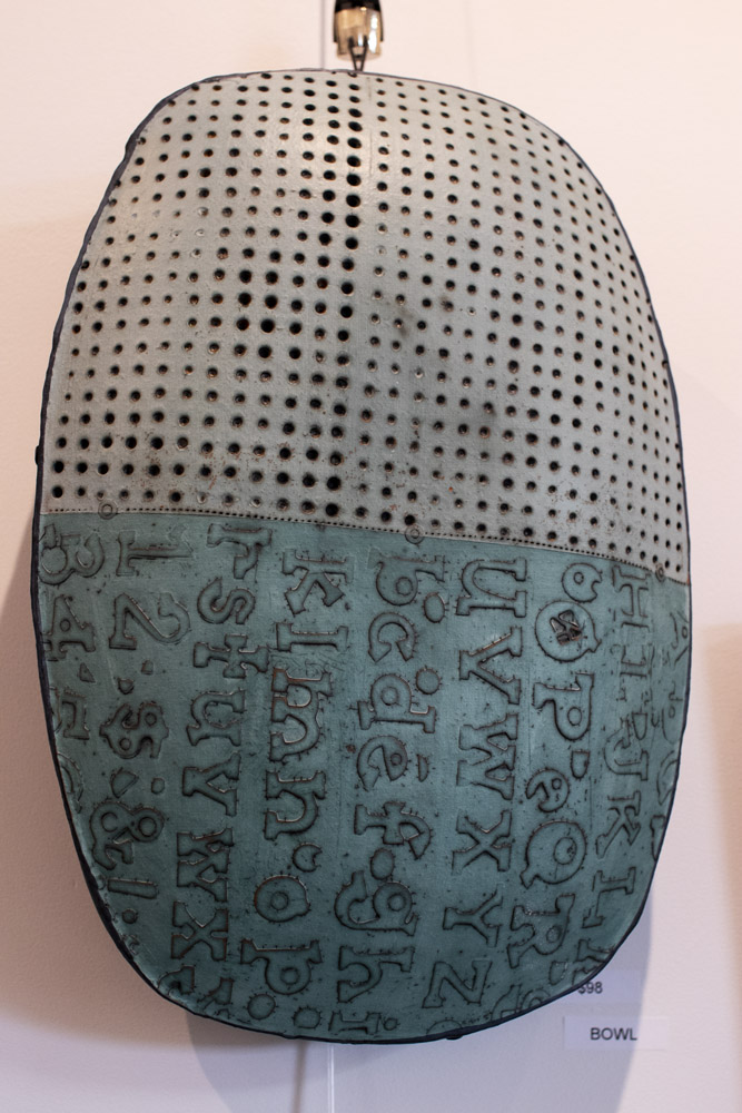 A white and teal pottery sculpture wall hanging is mounted to a white wall. The pottery is an oval shape with slightly square edges. The top half is white with deep circles carved into the clay in a grid layout. The bottom half is a deep teal with random letters carved in. This pottery is by Ann Ohotto Thompson for County 8 Pottery.