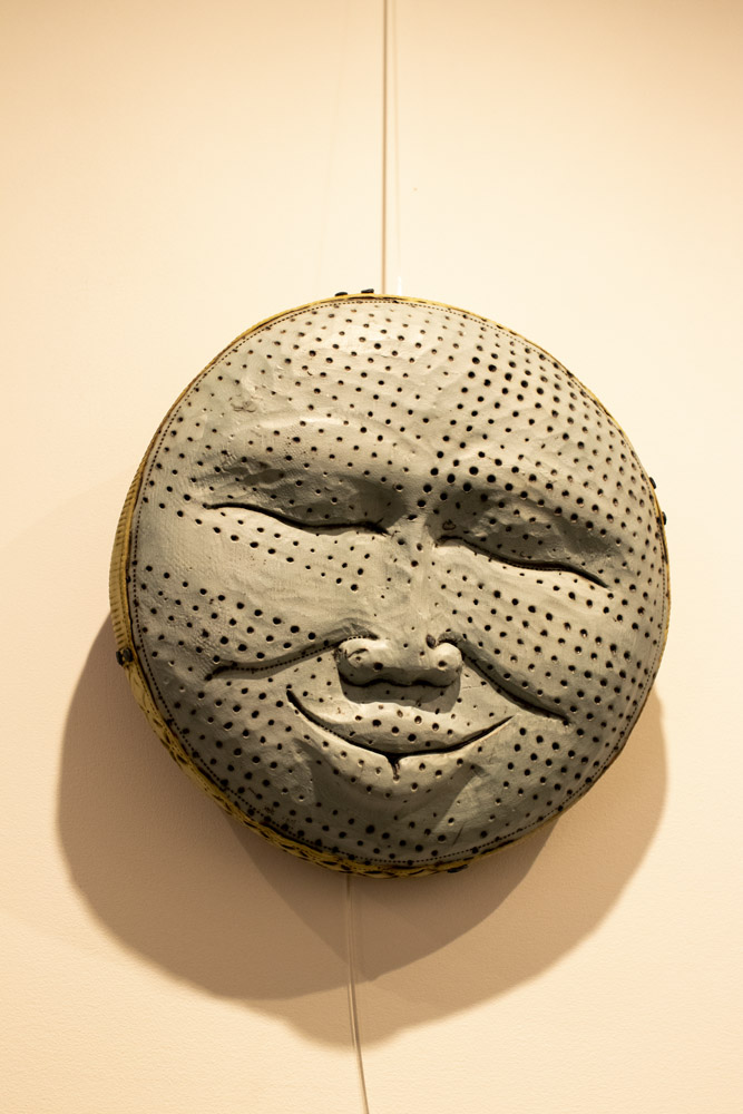 Photo shows a pottery wall hanging sculpture against a white wall. It is a big circle showing a face "Man in the Moon" design. The face has his eyes closed and shows a slight smile. There are circles textured all over the design like craters of the moon. The sculpture is one color grey all over. Pottery is by Ann Ohotto Thompson of County 8 Pottery.