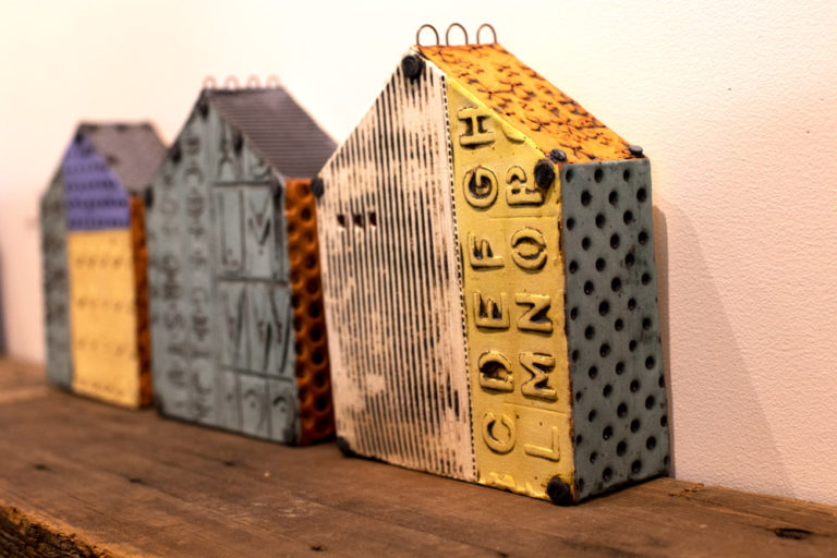 Three abstract house shaped pottery decorative wall hangings or sculptures sit on a dark wooden shelf against a white wall. They are decorated with deep contrasting textures like lines, letters, or circles, and glazed with a variety of colors like white, yellow, and light blue. All pottery is handmade by Ann Ohotto Thompson for County 8 Pottery.